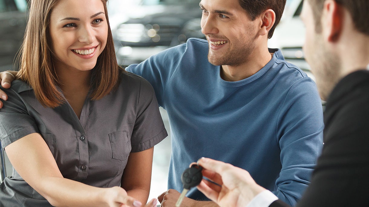 Woman reaches out to accept the keys from a sales person while the man sitting next to her has his arm around her shoulder smiling.