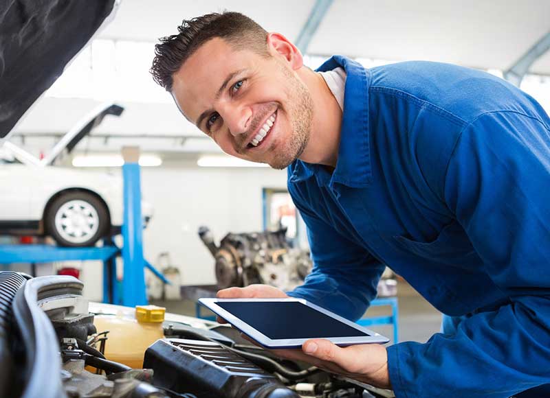 Service tech smiles at camera while holding a tablet and leaning over open engine bay